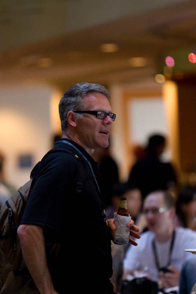 Ein Mann mit Rucksack, Brille und einem Ausweis hält eine Flasche, mit einem unscharfen Hintergrund.