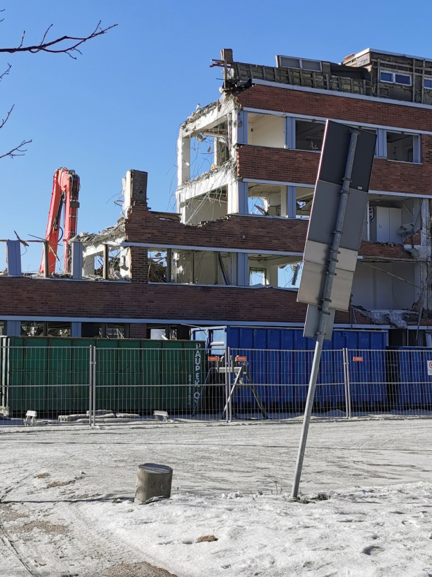 Großes Gebäude in einem Parkplatz abgerissen, umgeben von einem Metallzaun, Container, Pfählen, Schild, Baum und bewölktem Himmel.