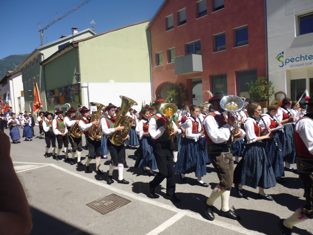 Eine Gruppe von Menschen in traditioneller bayrischer Tracht, die Musikinstrumente spielen, während sie eine Straße mit Gebäuden entlanggehen, einige halten Fahnen, mit einem Hügel und blauem Himmel im Hintergrund.