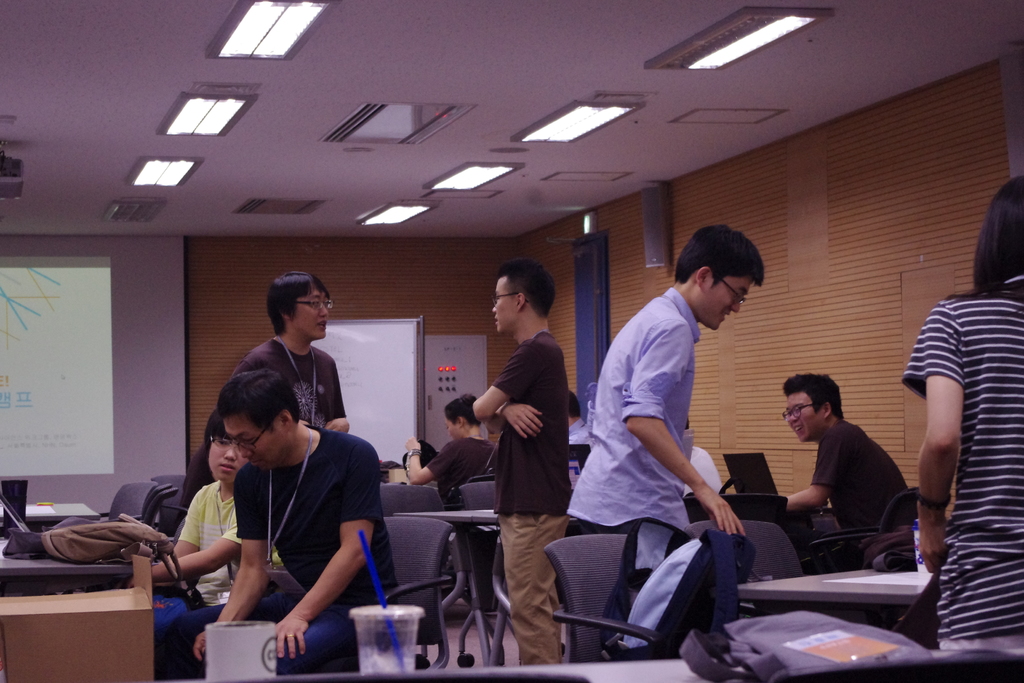 Ein Klassenzimmer mit Tischen, Stühlen, Laptops und Taschen, Menschen, die stehen und sprechen, ein Projektionsschirm und eine Holzwand im Hintergrund.