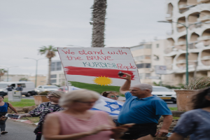 Eine Gruppe von Menschen marschiert nachts eine Straße entlang und hält ein Schild hoch, auf dem "Wir stehen zu den tapferen Kurdischen Menschen" steht, mit einem Wagen, der am Straßenrand geparkt ist, Straßenlaternen, Bäumen, Gebäuden und einem klaren blauen Himmel im Hintergrund.