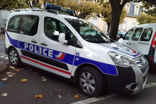 Ein Polizeiwagen steht am Straßenrand geparkt, mit Bäumen und Gebäuden im Hintergrund und einem sichtbaren Vorderrad.