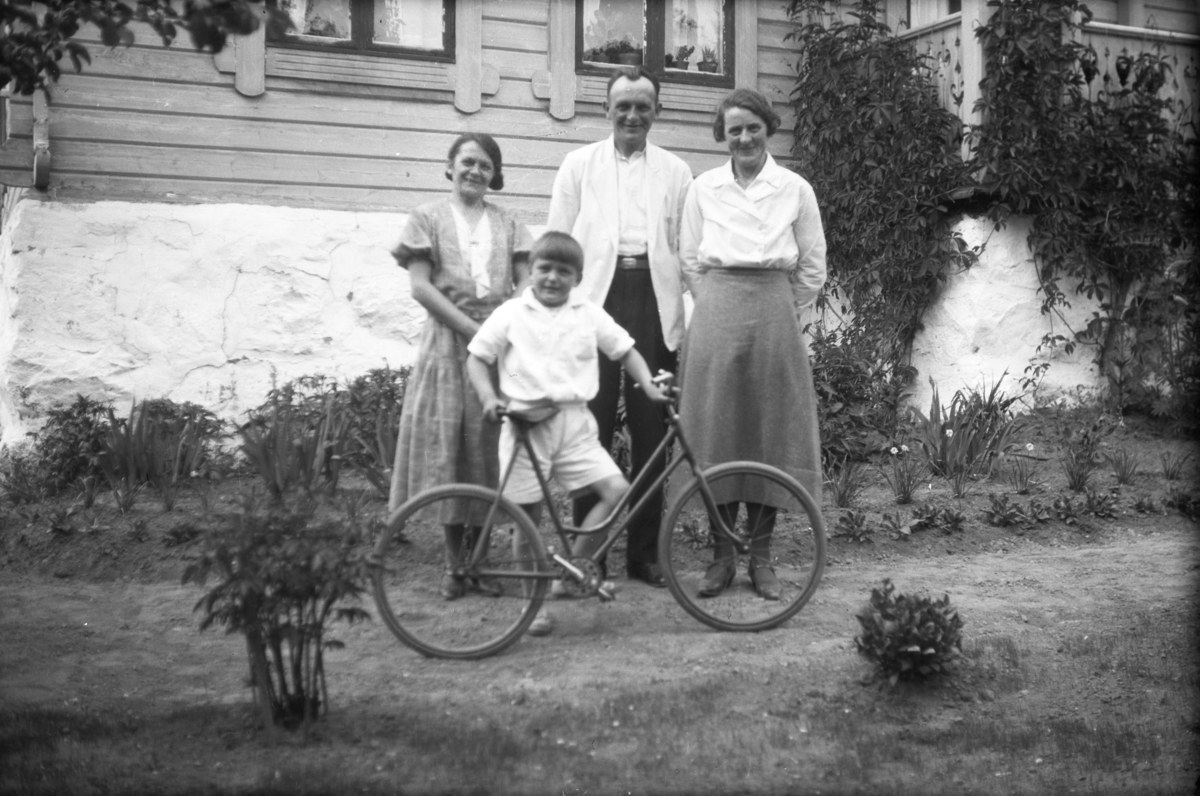 Schwarzes und weißes Foto einer Familie, die mit einem Jungen auf einem Fahrrad posiert, umgeben von Pflanzen, Gras, einem Gebäude und einem Baum.