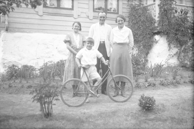 Schwarzes und weißes Foto einer Familie, die mit einem Jungen auf einem Fahrrad posiert, umgeben von Pflanzen, Gras, einem Gebäude und einem Baum.