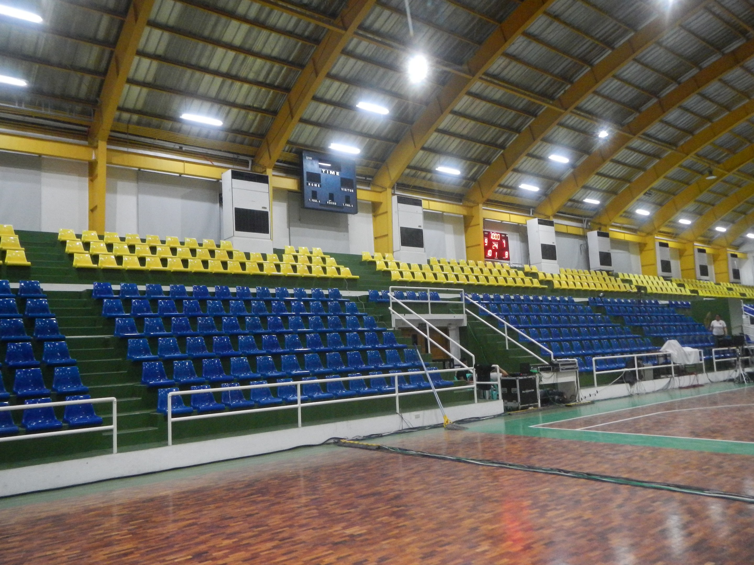 Indoor-Sporthalle mit Holzboden, blauen und gelben Sitzplätzen, Geländern, Treppen, wandmontierten Bildschirmen, Deckenleuchten und einigen Menschen.