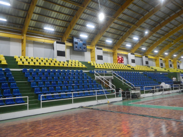 Indoor-Sporthalle mit Holzboden, blauen und gelben Sitzplätzen, Geländern, Treppen, wandmontierten Bildschirmen, Deckenleuchten und einigen Menschen.