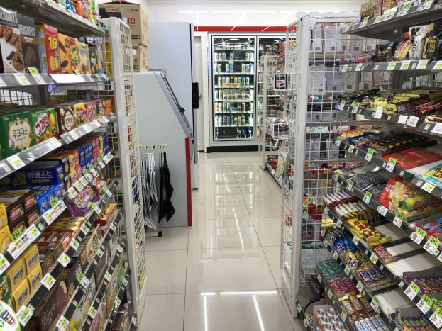 Ein Supermarktgang mit verschiedenen Lebensmitteln auf Regalen und Ablagen, mit Preisschildern, einem Kühlschrank im Hintergrund und Deckenbeleuchtung.