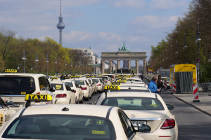 Eine belebte Straße in Berlin, Deutschland, mit zahlreichen parkenden Taxis, Fußgängern auf dem Gehweg, Laternen, Bäumen und Gebäuden im Hintergrund, mit einem fernen Bogen mit Statuen und einem Turm unter einem bewölkten Himmel.