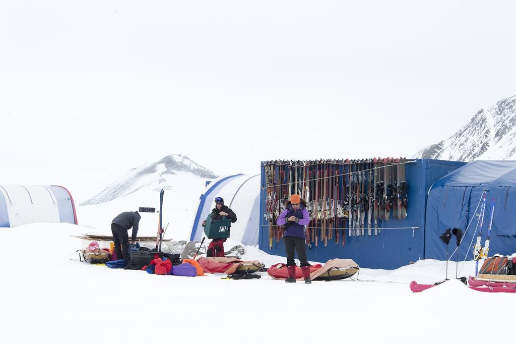 Drei Personen stehen auf einer schneebedeckten Landschaft mit verstreuten Taschen, Zelte mit Skiern dahinter und schneebedeckten Hügeln im Hintergrund unter einem klaren Himmel.