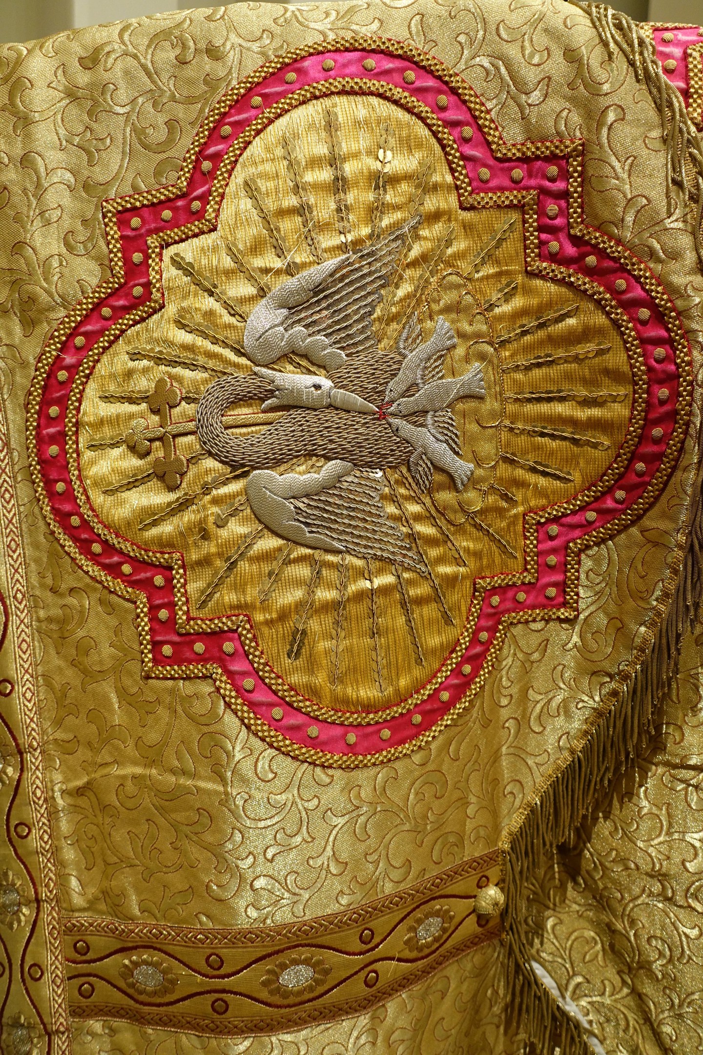 Nahaufnahme eines Priestergewandes mit aufwendiger goldener und roter Stickerei vor einem neutralen Wandhintergrund.