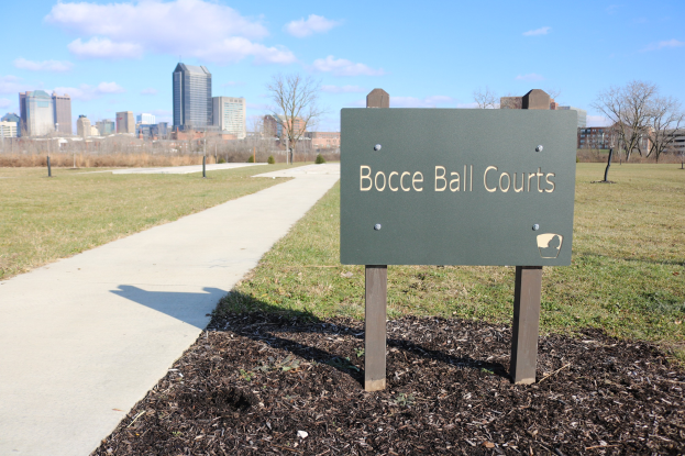 Ein Schild am Boden neben einem Weg und Bäumen trägt die Aufschrift "Bocce Ball Courts in Indianapolis, Indiana" mit einem bewölkten Himmel darüber.