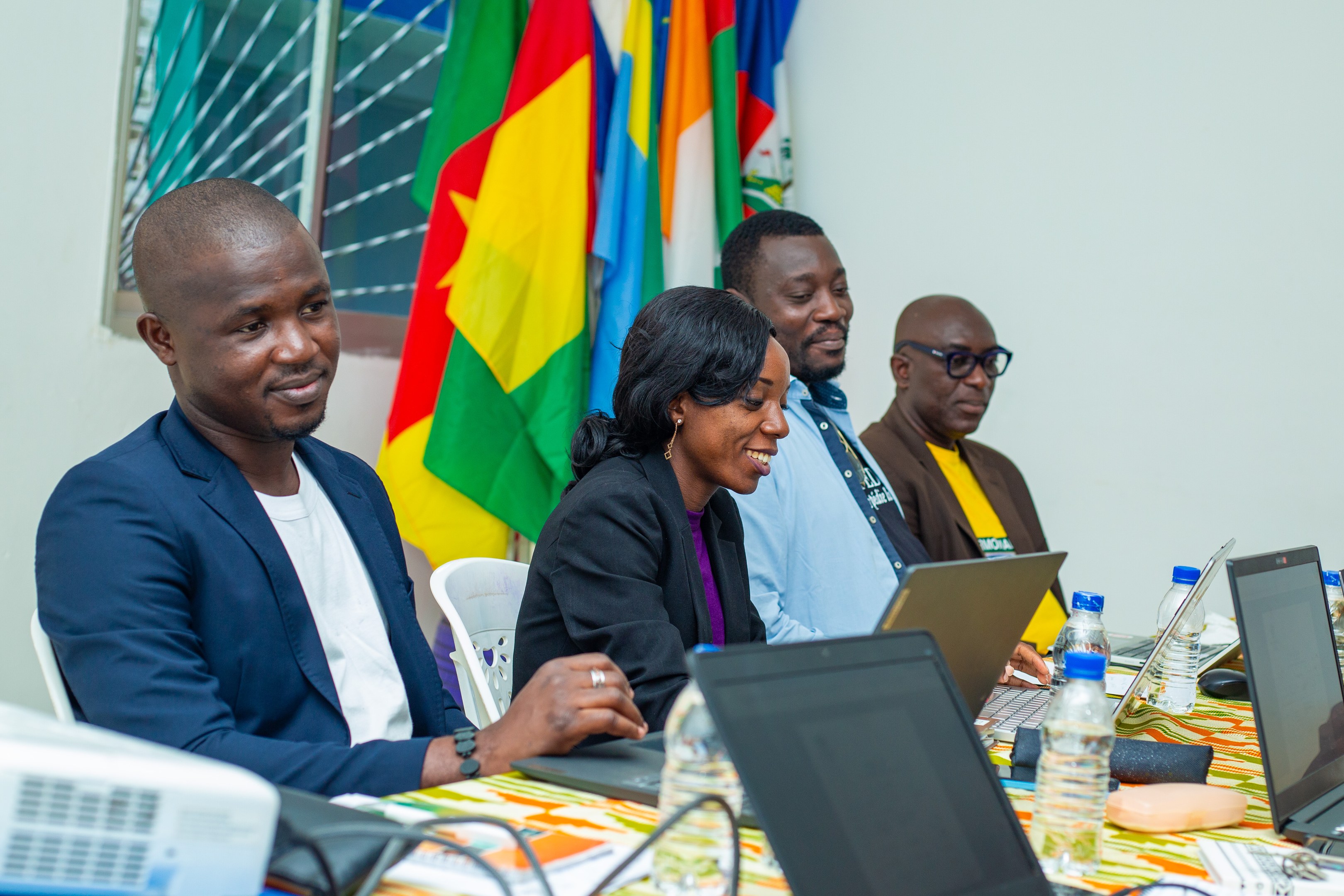Eine Gruppe von Menschen in einem Konferenzraum mit Laptops, Flaschen, Mikrofonen und Papieren, die sich in einer Diskussion mit ghanaischen Flaggen im Hintergrund befinden.