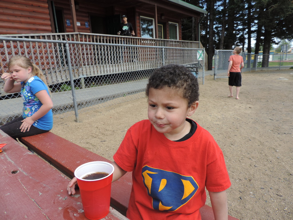 Ein Kind sitzt auf einer Bank mit einem Glas Getränk vor sich, ein Mädchen neben ihm und ein Junge, der hinter ihnen geht, mit einem Metallzaun, einem Haus mit einer Person und Bäumen im Hintergrund.