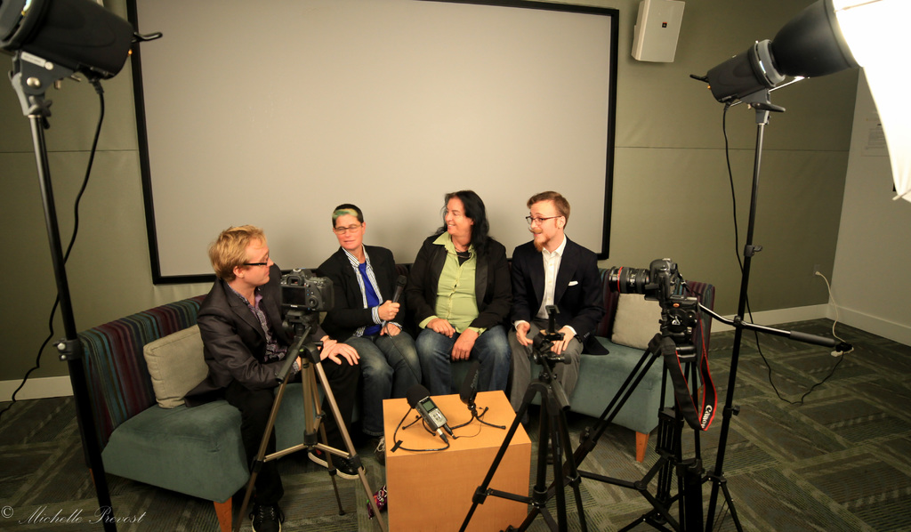 Eine Gruppe von drei Personen sitzt auf einem Sofa und gibt ein Interview, wobei eine Person links in einem schwarzen Mantel und Hose, eine Person in der Mitte mit einem Mikrofon haltenden Mantel und Krawatte und eine Person rechts mit einem Lächeln zu sehen ist. Ein Mikrofonrekorder befindet sich auf dem Mitteltisch, Kameras sind auf der rechten und linken Seite und große Scheinwerfer sind vorhanden, sowie eine Projektionswand im Hintergrund.
