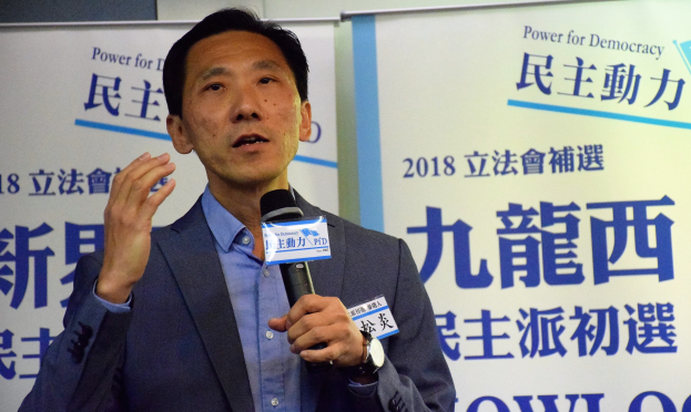 Ein Mann in Anzug und Krawatte spricht in ein Mikrofon an einem Podium mit Bannern im Hintergrund.