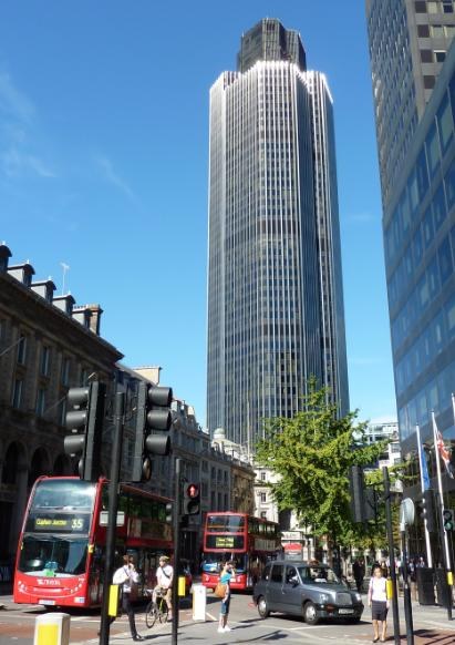 Eine belebte Stadtstraße in London mit Fahrzeugen, Fußgängern, Verkehrsampeln, Bäumen, Gebäuden, Flaggen und dem Tower der Bank of England im Hintergrund bei einem bewölkten Himmel.