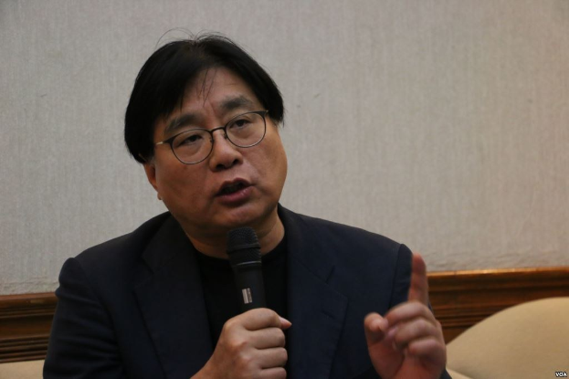 Ein Mann in Anzug und Brille spricht in ein Mikrofon und hält eine Rede vor einer Wand.