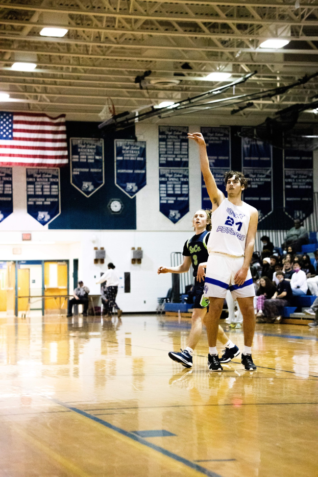 Mann in Sportbekleidung schießt Basketball in einer Turnhalle, während Zuschauer von Bänken im Hintergrund zusehen.