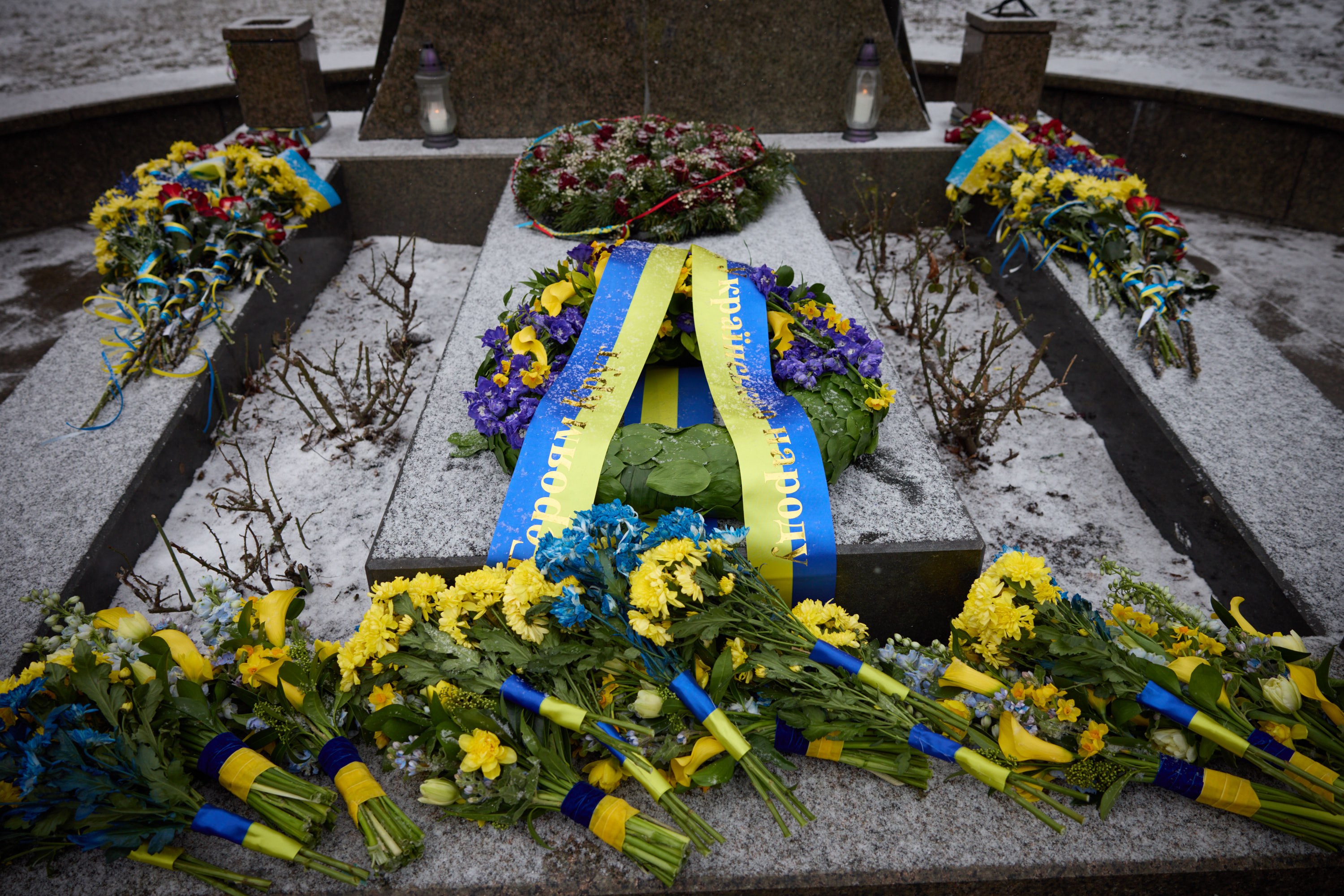 Gedenkstätte in Kiew, Ukraine, mit einem Blumenkranz auf einem Grab, das mit einem blauen Band markiert ist, umgeben von zusätzlichen Buketts, vor einem schneebedeckten Hintergrund.