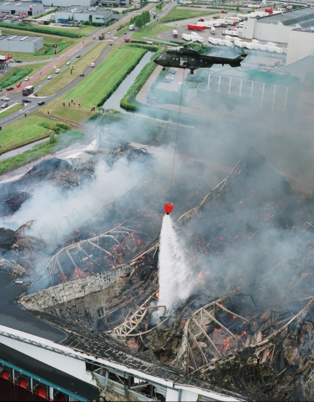 Ein Hubschrauber fliegt über ein großes, durch ein Feuer zerstörtes Gebäude, das Rauch ausstößt, mit mehreren Gebäuden, Fahrzeugen und Bäumen im Hintergrund und Text am unteren Rand.