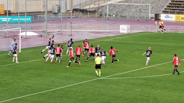 Gruppe von Menschen, die Fußball auf einem Feld mit Zuschauern in einem Stadion spielen, umgeben von Pfosten, Tafeln mit Text, Geländern und anderen Gegenständen.