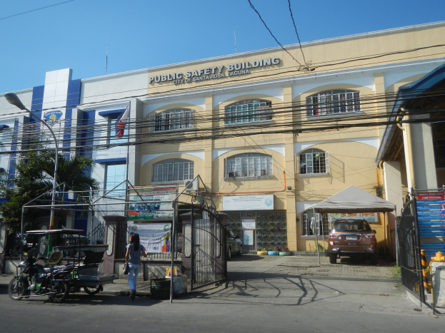 Gebäude für öffentliche Sicherheit umgeben von Bäumen, Straßeninfrastruktur, Fahrzeugen, Fußgängern und einem klaren Himmel.