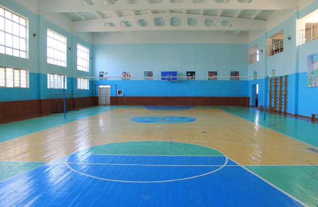 Großer Turnsaal mit blau-weißem Boden, Basketballfeld, Netz, Stangen, Tür, Fenstern, Wandplakaten, Geländer und Dach.