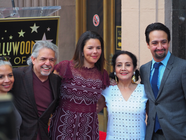 Gruppe von Menschen auf einem roten Teppich bei der Zeremonie des Hollywood Walk of Fame, mit einer Kamera auf der linken Seite, einer Tafel mit Text, einem Glaspodest und einer Wand im Hintergrund.