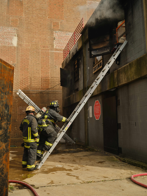 Feuerwehrleute mit Helmen klettern an einer Leiter zu einem raucherfüllten Gebäude, während in der Nähe ein metallisches Objekt und Bodenrohre zu sehen sind.