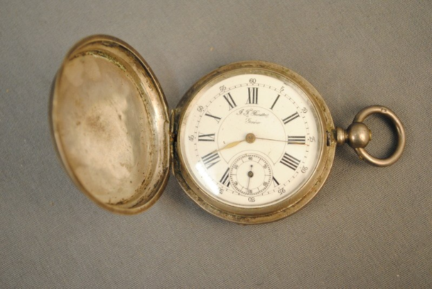 Eine silberne Taschenuhr mit einem weißen Zifferblatt, das römische Ziffern auf einem grauen Hintergrund zeigt, mit einem Deckel für einen klassischen Look.