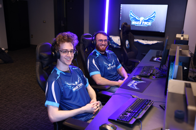 Zwei Männer mit Headsets sitzen an einem Schreibtisch mit Monitoren, Tastaturen und anderem Equipment und lächeln, während sie ein Spiel der Wake Tech League of Legends spielen zu scheinen.