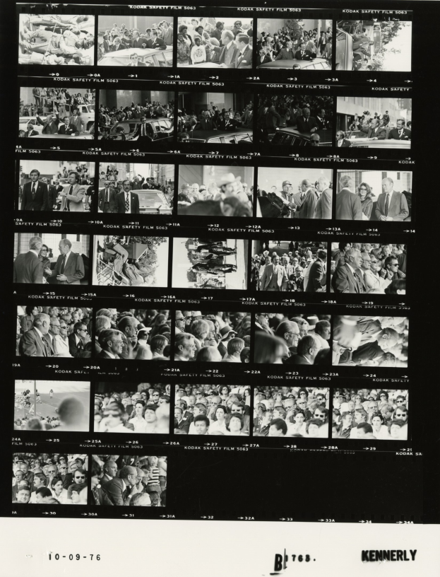 Schwarzes und weißes Foto einer vielseitigen Menschenmenge, die fröhlich in die Kamera schaut, während sie auf einen Fernsehbildschirm mit Text am unteren Rand schaut.