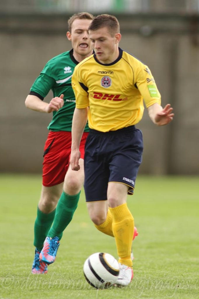 Zwei Männer spielen Fußball auf einem grünen Feld mit einem Ball, mit unscharfem Hintergrund und Text unten.