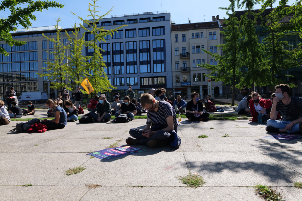 Eine Gruppe von Menschen sitzt vor einem Gebäude auf dem Boden, einige tragen Masken, mit verstreuten Taschen und Gegenständen, während einer Demonstration in Berlin bei klarem blauem Himmel.