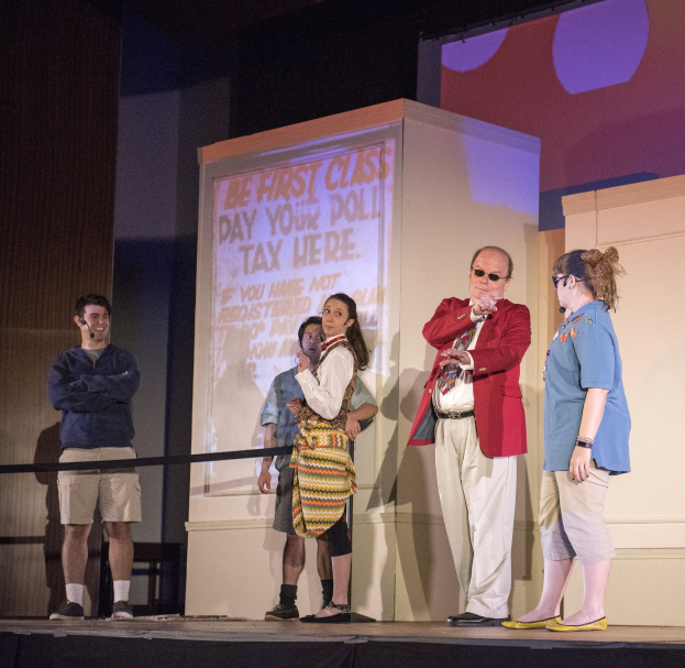 Eine Gruppe von Menschen auf einer Bühne mit einem Mann, der ein Mikrofon hält, einem Bildschirm dahinter mit Text, einem Zaun, einer Wand und Deckenlampen.