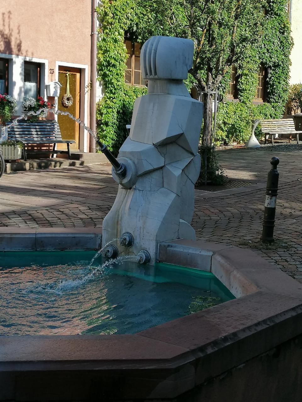 Ein steinerner Brunnen mit fließendem Wasser in der Mitte eines Stadtplatzes, umgeben von Gebäuden, Grünflächen, Bänken und Blumenkübeln.