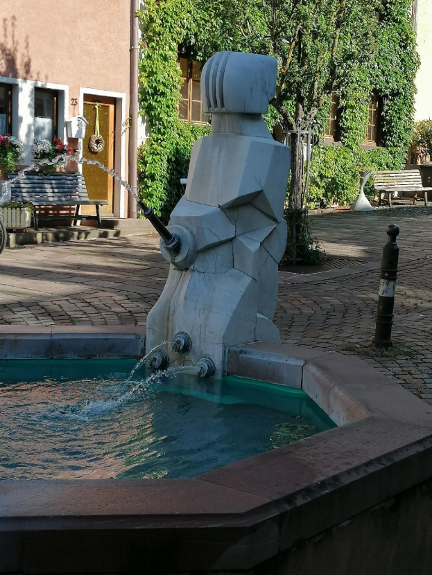 Ein steinerner Brunnen mit fließendem Wasser in der Mitte eines Stadtplatzes, umgeben von Gebäuden, Grünflächen, Bänken und Blumenkübeln.