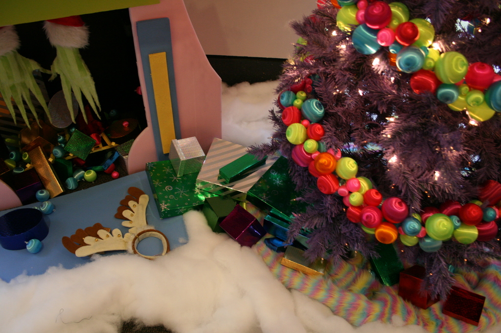 Eine Indoor-Szene mit einem Weihnachtsbaum mit Lichterketten und Dekorationen auf der rechten Seite, Geschenkboxen und Dekorationsgegenständen in der Mitte und einer Hütte mit Dekorationen und Watte in der oberen linken Ecke, mit einer Wand im Hintergrund und Watte und wollener Stoff unten.