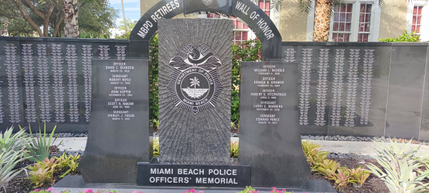 Miami Beach Police Officers Memorial mit einem zentralen Stein, umgeben von Grün, Bäumen im Hintergrund und einem Gebäude unter einem sichtbaren Himmel.