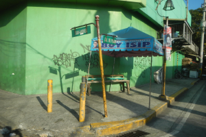 Ein grünes Gebäude mit Graffiti, einem blauen Zelt, einer Bank, Pfosten, Lampen, Schildern, einem Fahrrad, Bäumen und anderen Gegenständen im Vordergrund und einem Himmel im Hintergrund.