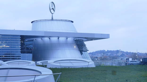 Großes modernes Mercedes-Benz-Museum-Gebäude mit Logo oben, umgeben von Gras, Geländern, Fahrzeugen auf der Straße, Bäumen und einem klaren blauen Himmel.