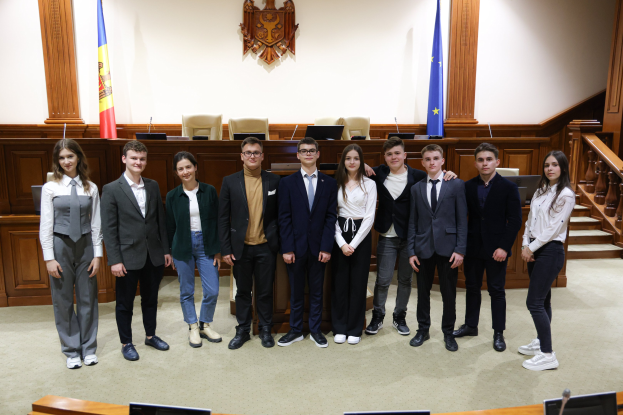 Gruppe von lachenden Menschen vor einem Gerichtssaal mit Monitoren und Mikrofonen auf Tischen, einer hölzernen Plattform mit Fahnen und Stühlen im Hintergrund und einer Treppe auf der rechten Seite.