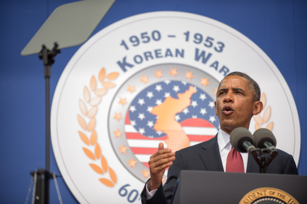 Präsident Obama steht an einem Rednerpult, trägt ein Sakko, Krawatte und Hemd und hält eine Rede am Korean War Memorial in Washington, D.C., mit einem Banner und Emblem dahinter und einem Ständer links daneben.