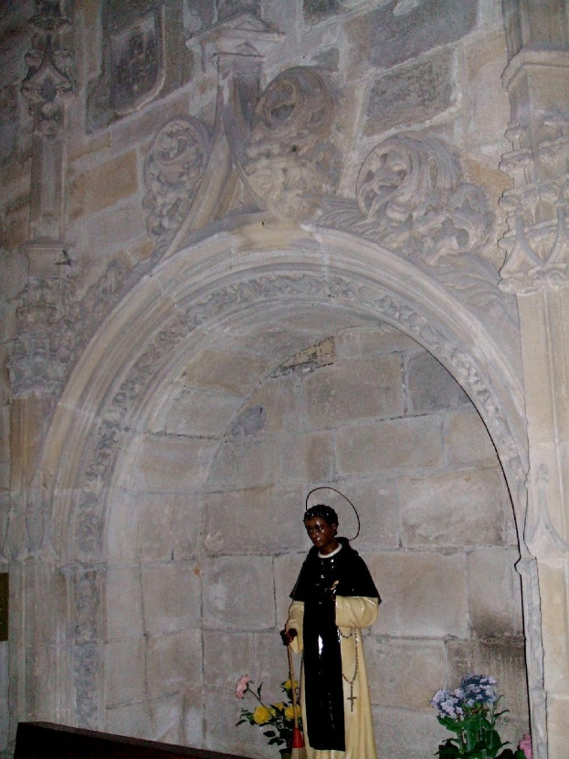 Ein Priester in schwarzer und cremefarbener Robe steht in einem Kirchenbogen und hält einen Stab, mit blühenden Pflanzen neben ihm und kunstvoll verzierten Wänden dahinter.