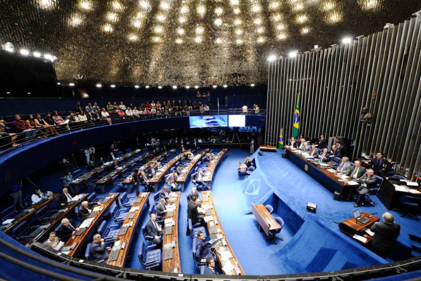 Eine Konferenzbühne mit sitzenden Personen an einem Tisch mit Papieren und Gegenständen, flankiert von Fahnen und einer Skulptur, mit Zuschauern an Tischen und hinter einer Absperrung, unter dekorativen Deckenleuchten und zwei Bildschirmen.