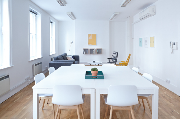 Innenansicht eines Apartments in Berlin mit einem weißen Tisch mit einer Pflanze in einer Schale, Stühlen, einer Couch mit Kissen, einer Lampe, Regalen mit Gegenständen, einer Klimaanlage, Fotorahmen an der Wand, Fenstern und Deckenleuchten.