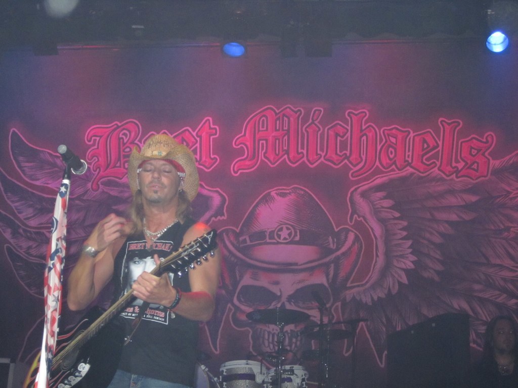 Ein Konzertbild mit einem Gitarristen links neben einem Mikrofon, einer Person in der rechten unteren Ecke, einem Banner im Hintergrund und Drogen in der Mitte unten, mit Scheinwerfern oben.