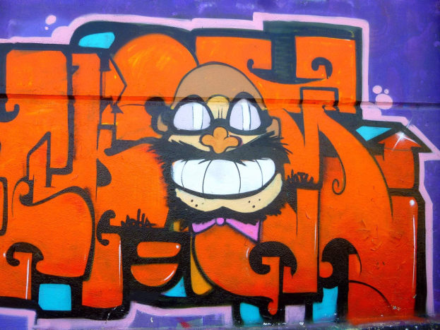 Graffiti-Gemälde eines Cartoon-Charakters mit großen Augen, einem breiten Lächeln und einem verschmitzten Ausdruck an der Seite eines Gebäudes, mit leuchtenden Farben und auffälliger Begleittext.