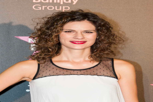 Eine Frau in einem schwarzen Kleid mit tiefem Ausschnitt und schwarzen High Heels, die auf einem roten Teppich steht, strahlt und posiert für die Kamera, mit einem Banner im Hintergrund mit der Aufschrift "Maggie Gyllenhaal bei den Billboard Music Awards 2016".