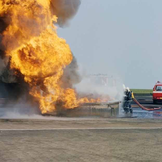 Löschfahrzeug in Flammen an der Seite einer Straße mit zwei Helmet tragenden Individuen, die Rohre halten, einem Fahrzeug im Hintergrund und dem Himmel.